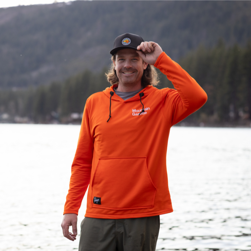 Male model wearing a Mountain Gazette hat and the Mountain Gazette x Burgeon Outdoor Hooded Sweatshirt in Orange by a lake
