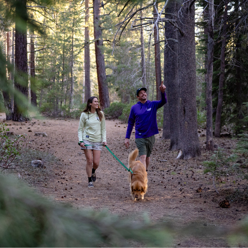 Male and female models wearing the Mountain Gazette x Burgeon Outdoor Hooded Sweatshirt while walking a dog in the woods