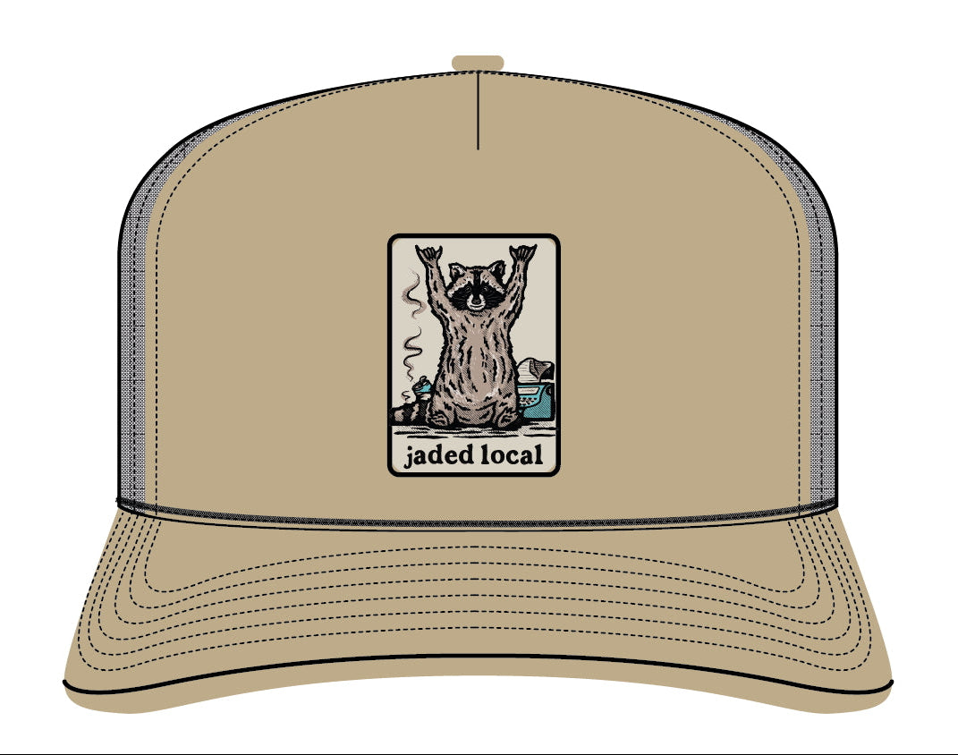 A tan trucker hat with a front graphic patch showing a raccoon and the text 'jaded local'. The hat has a tan front panel and brim, dark gray mesh back panels, and black stitching.