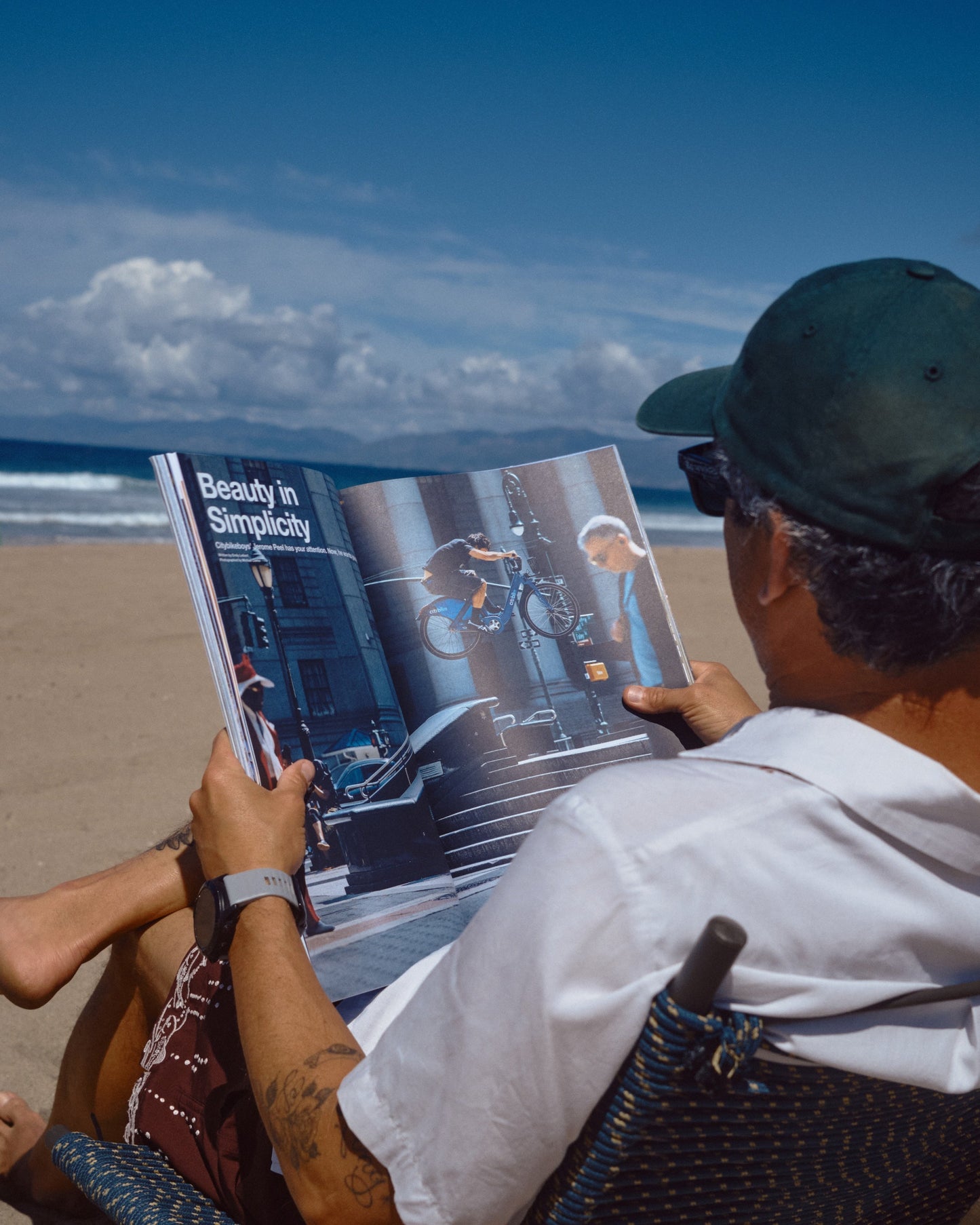 Person reading Mountain Gazette 205 magazine on a beach with ocean and sky in the background