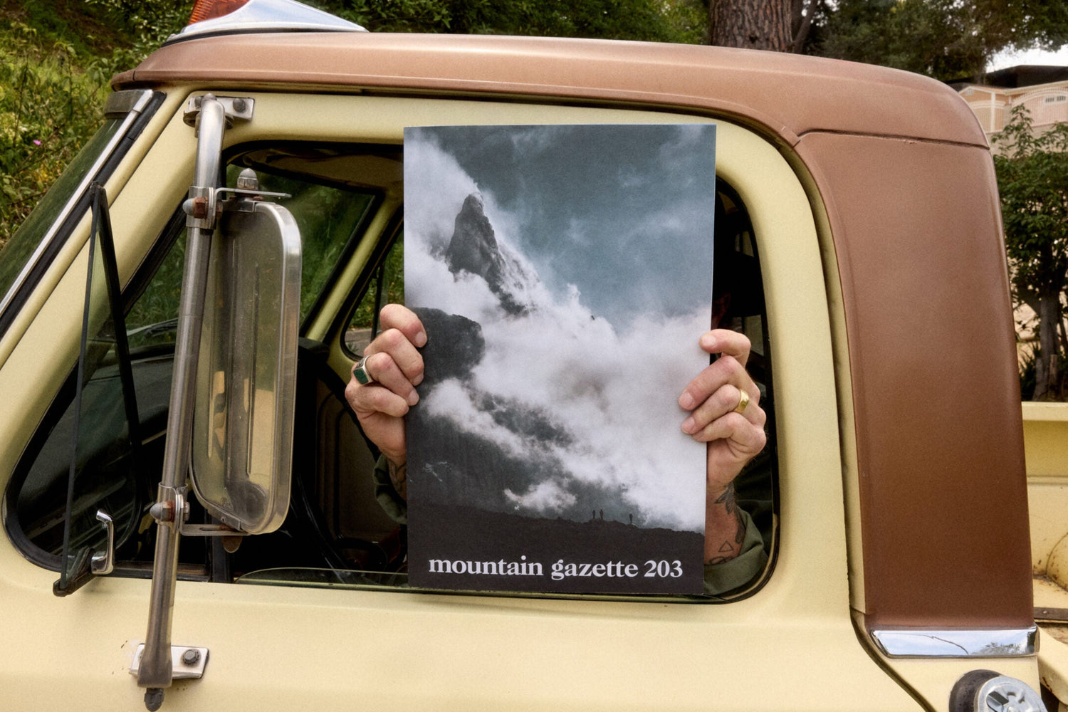 Person holding a magazine titled 'mountain gazette 203' in front of a vehicle window with a scenic background.