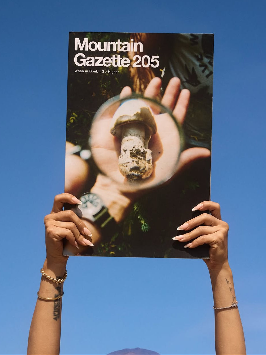 Person holding a magazine titled 'Mountain Gazette 205' against a clear blue sky