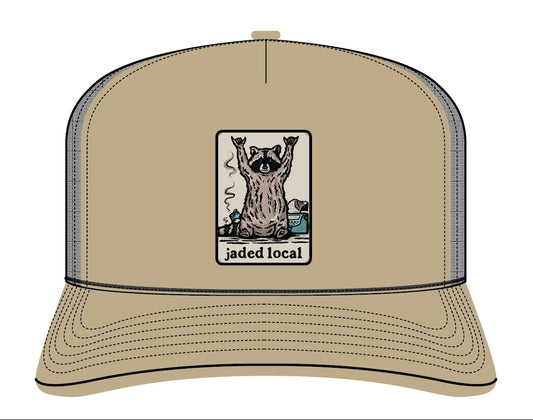 A tan trucker hat with a front graphic patch showing a raccoon and the text 'jaded local'. The hat has a tan front panel and brim, dark gray mesh back panels, and black stitching.