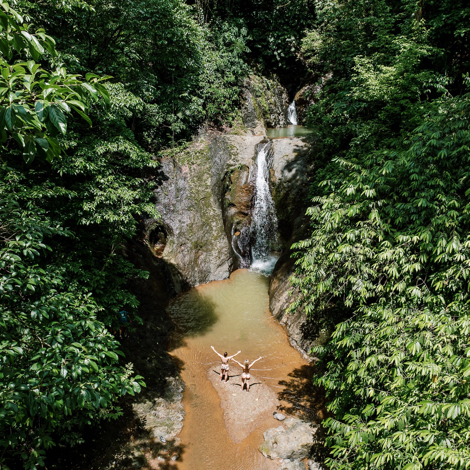 Two people standing in a river at the base of a waterfall surrounded by lush greenery.